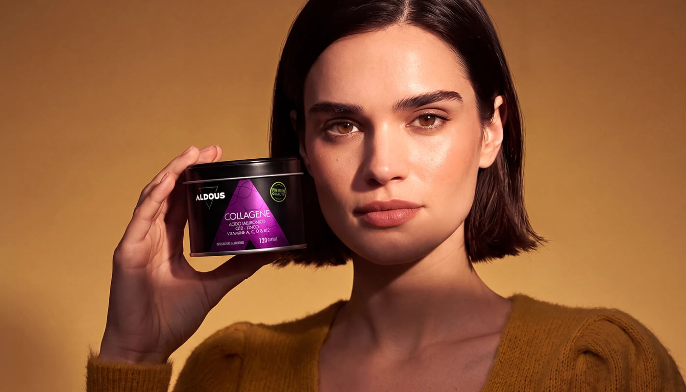 Una donna con capelli corti scuri e un maglione color senape mostra una confezione nera di integratore in capsule Aldous Collagene, posando davanti a uno sfondo dai toni gialli e arancioni.